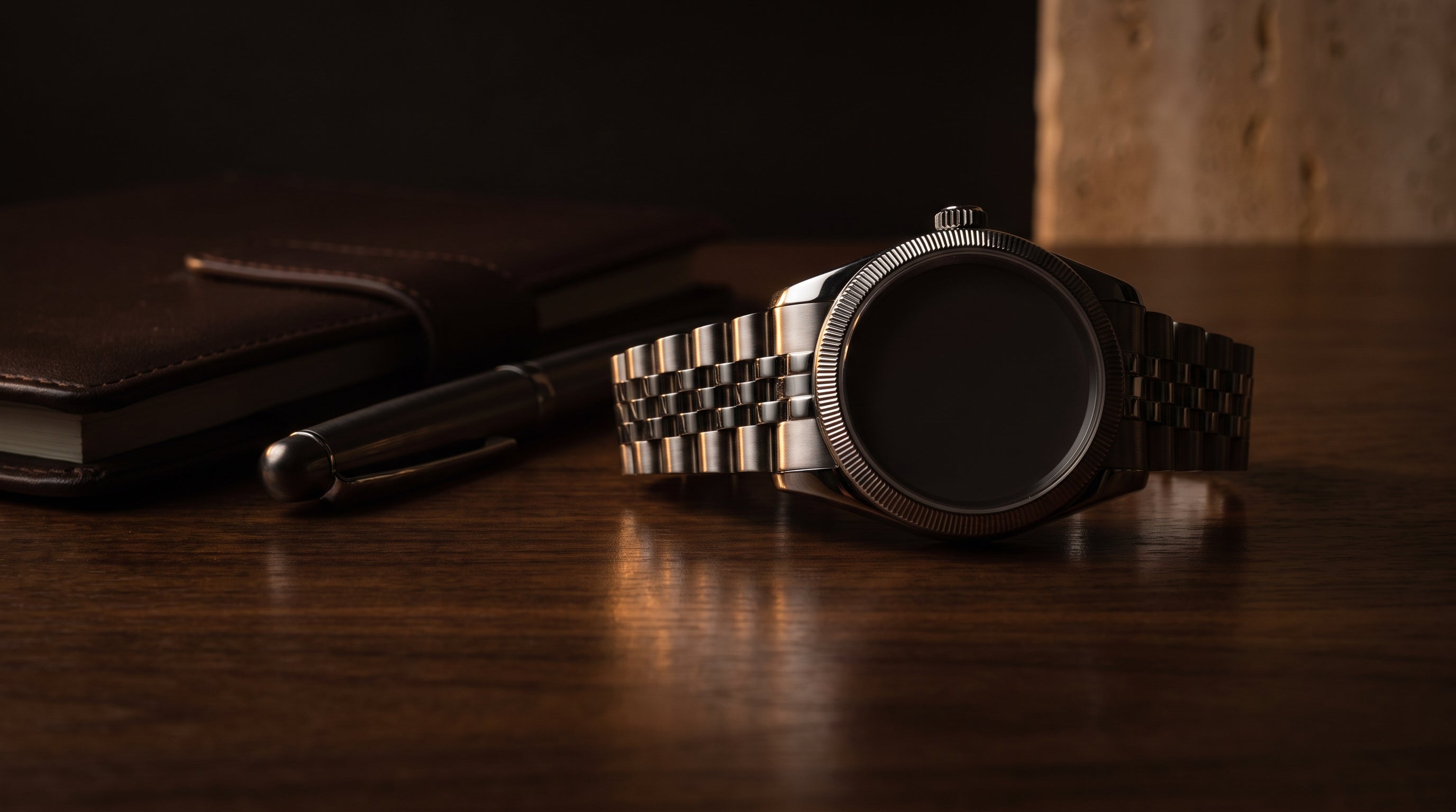 Steel dress watch with a fluted-style bezel and five-link bracelet on a dark walnut desk in warm, low-key editorial light